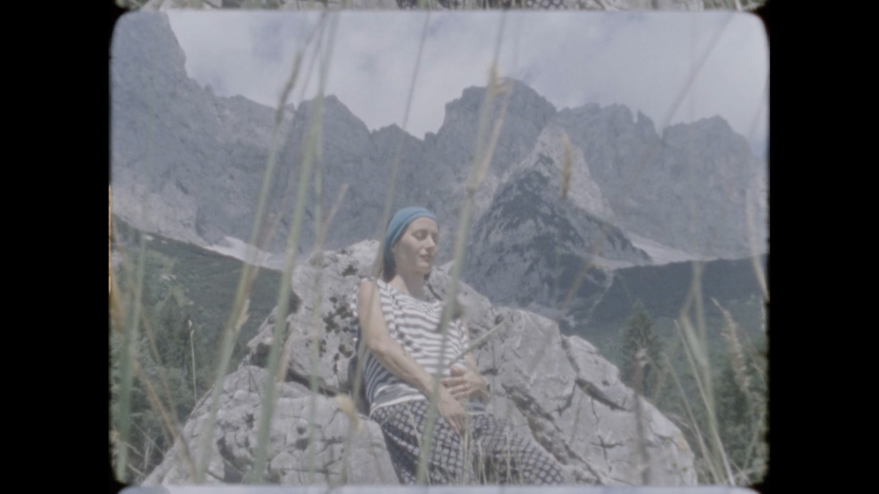 A woman sitting on a rock in the mountains, filmed through tall grass
